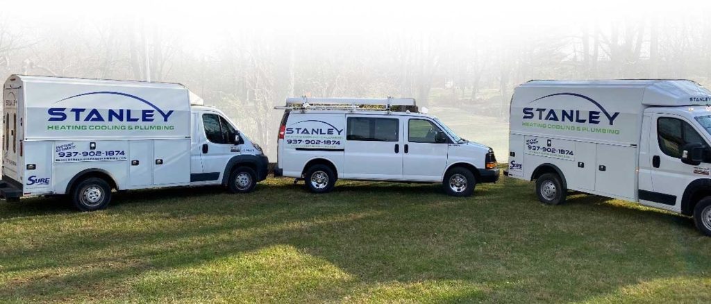 Three white Stanley Heating, Cooling & Plumbing service vans are parked on grass, facing slightly different directions. The company logo and phone number are visible on each vehicle. Trees and a light sky are in the background.