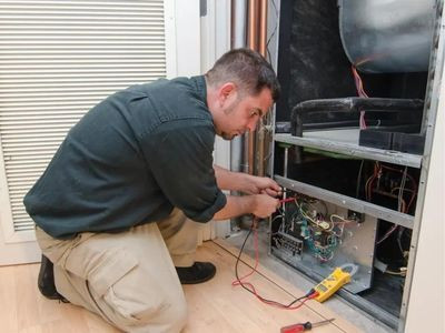 An HVAC technician in a black shirt and khaki pants kneels on the floor, using a multimeter to check wiring inside an open HVAC unit. Tools are scattered nearby—a typical scene showcasing the essential work of HVAC technicians we thank this Thanksgiving.