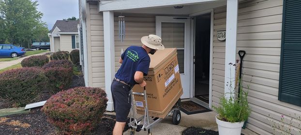 A person wearing a hat and navy shirt uses a hand truck to deliver a large boxed item to the front door of a house with beige siding and green shutters. Shrubs line the walkway leading to the entrance.