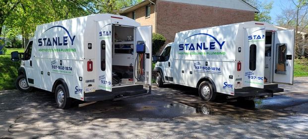 Two white Stanley Heating Cooling & Plumbing service vans are parked outdoors with their back doors open, revealing equipment inside. The vans are near a house on a sunny day with green trees in the background.