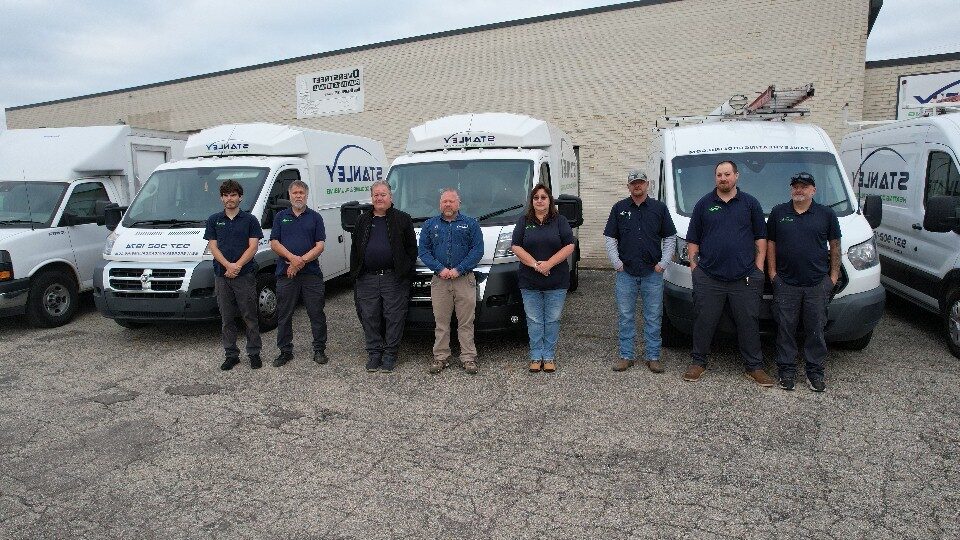 Eight people stand in a line in front of several white service vans parked on a cracked lot, all wearing dark clothing, with a brick building in the background.