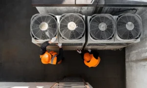 Two workers in orange safety vests and helmets inspect four large rooftop air conditioning units—an important part of learning How Long Do HVAC Systems Last—in an industrial setting, viewed from above.