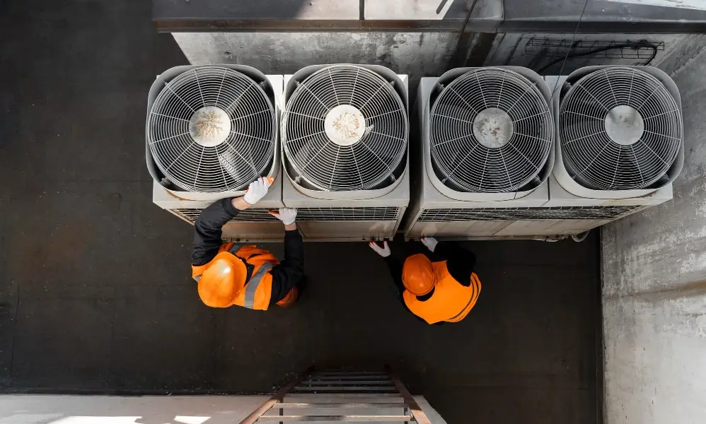 Two workers in orange vests and helmets inspect four large industrial air conditioning units from above, highlighting the importance of regular maintenance to determine how long HVAC systems last in an outdoor urban setting.