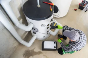 A technician wearing gloves and a cap is servicing a water heater, inspecting controls and pipe connections to address water heater odor, using tools on a clean, light-colored floor.