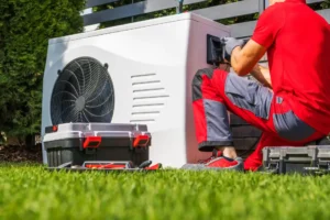 A technician in red work clothes kneels on grass while providing Heat Pump Repair for an outdoor unit, with toolboxes nearby.