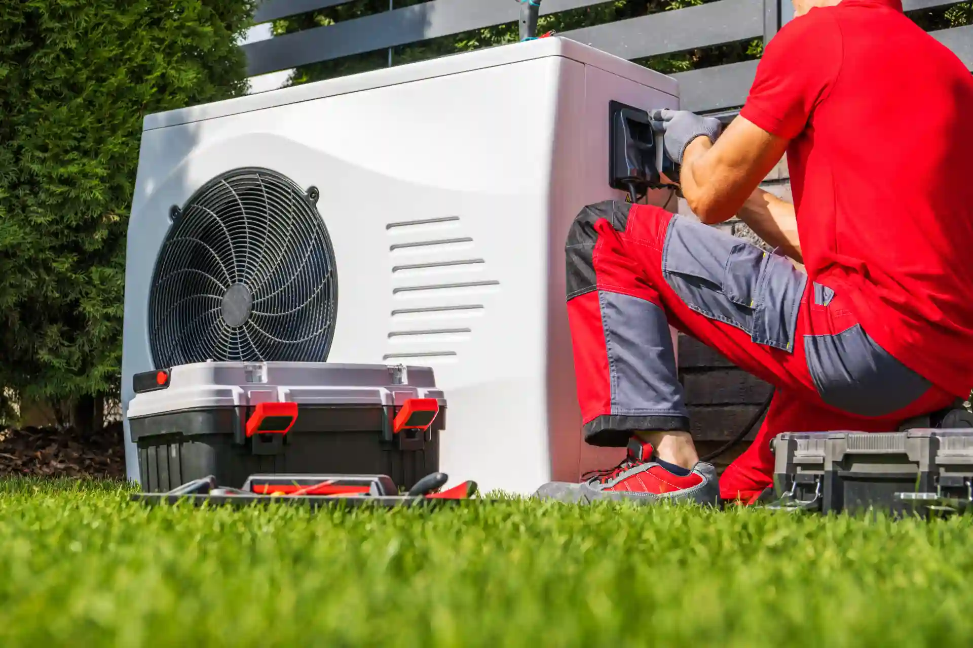 A technician in red work clothes kneels on grass while providing Heat Pump Repair for an outdoor unit, with toolboxes nearby.