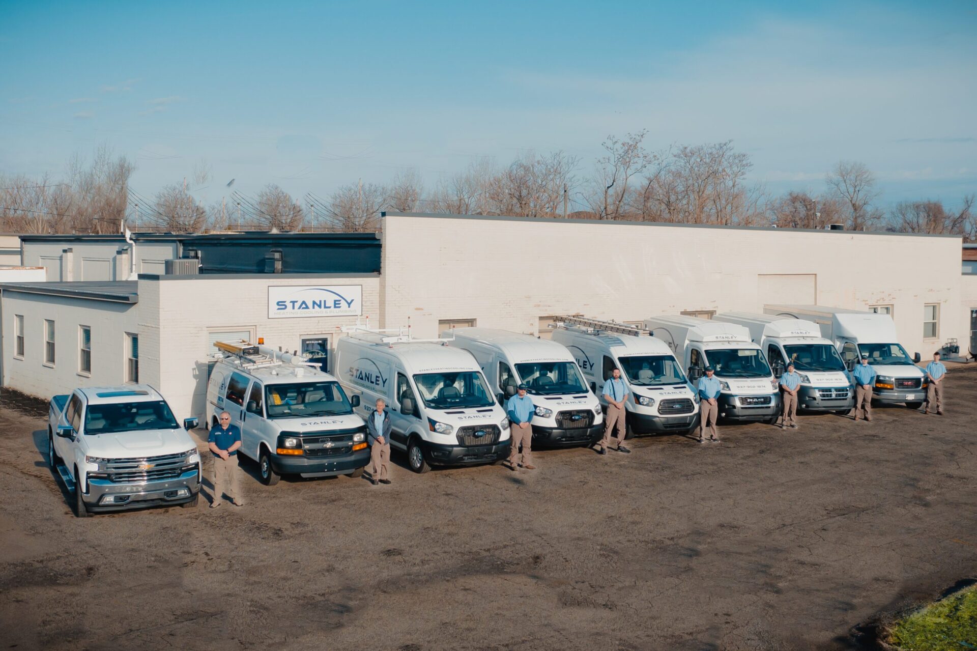 Eight people stand in front of a row of work vans and trucks parked outside a white industrial building with a Stanley sign, under a clear blue sky.