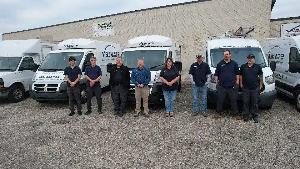 Eight people stand in a row in front of several Stanley HVAC repair vans, posing for a group photo in a parking lot next to a light-colored brick building.