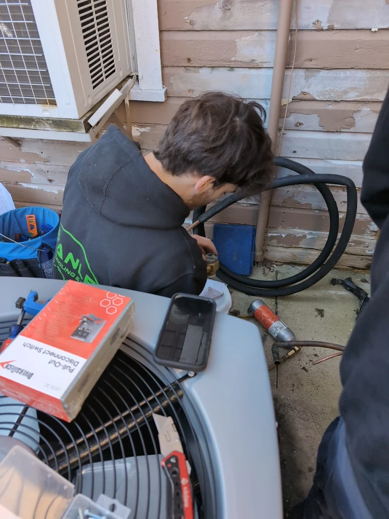 A person kneels on the ground outside in Moraine, working with tools and pipes near an air conditioning unit—perhaps handling a summer AC repair cost. A phone, box, and utility knife rest on top of the unit; behind, the wall’s paint is peeling.