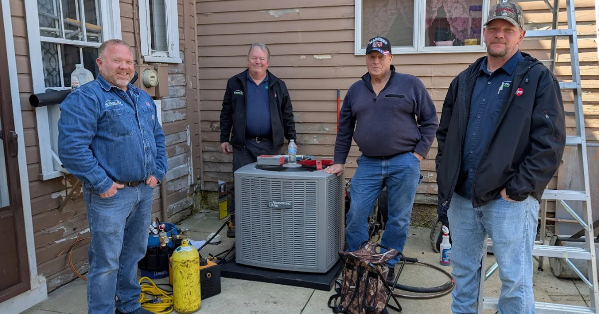 Four men stand outside a house next to a newly installed air conditioning unit, with tools and equipment visible nearby. The siding of the house shows signs of wear, and a ladder leans against the wall.