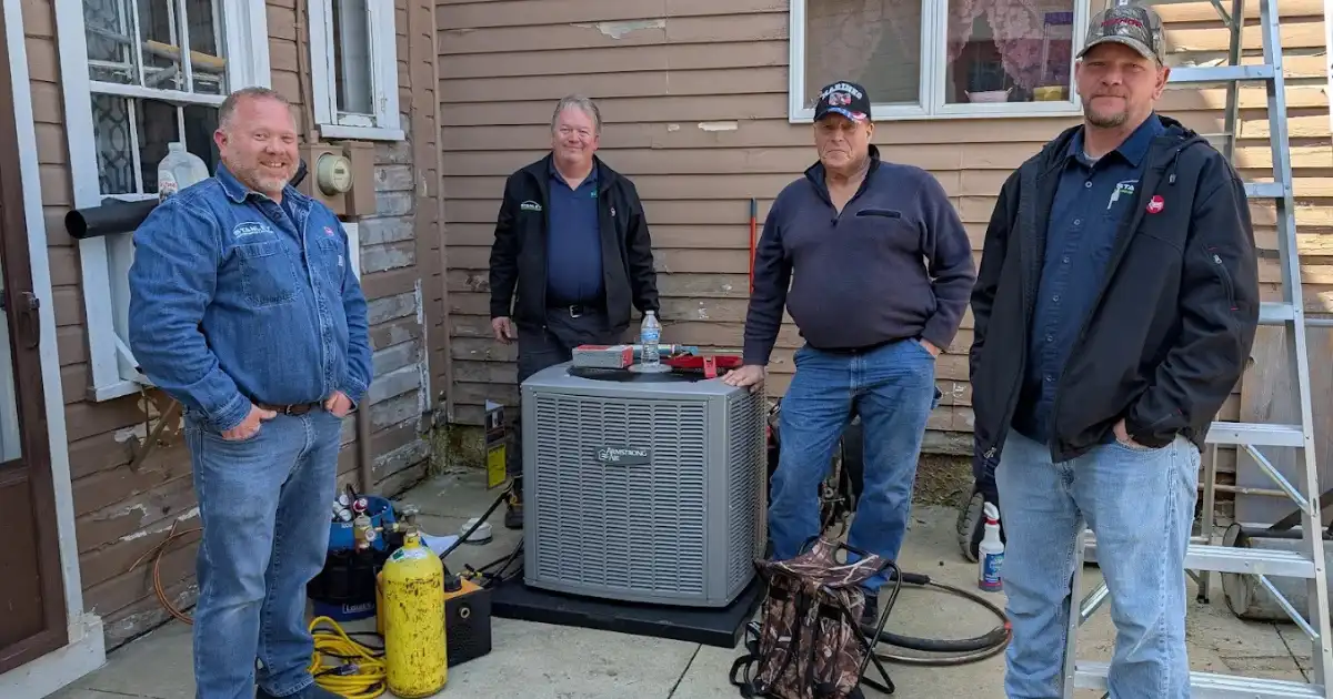 Four men stand outside a house in Dayton OH near a new air conditioning unit and various tools. The house siding is partly repaired, with a metal ladder on the right. They appear ready to handle any emergency heating repair needs.