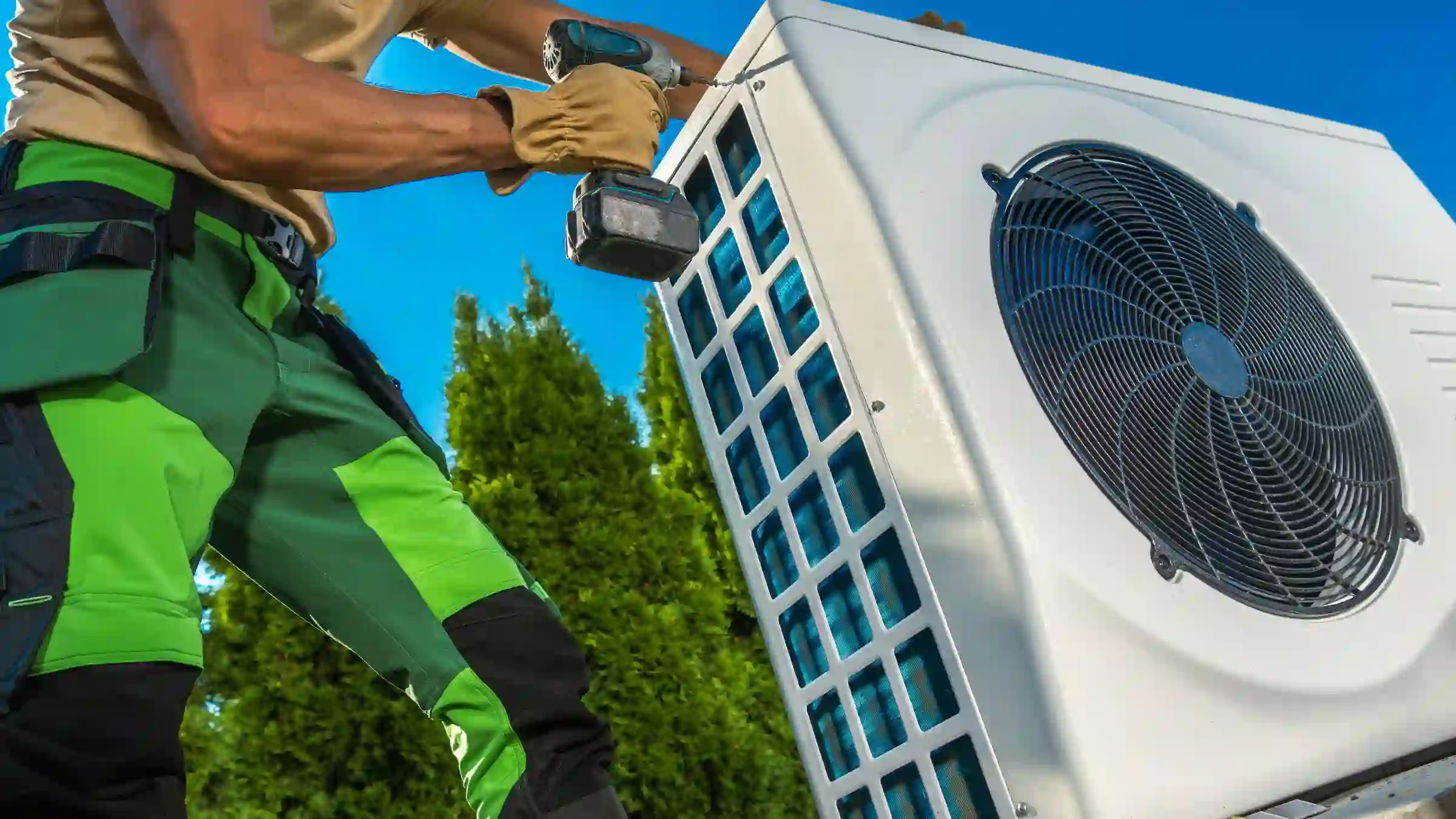 A worker in green and black pants uses a power drill to repair an air conditioner outdoors, with trees and a clear blue sky in the background—a typical scene during professional air conditioner repair.