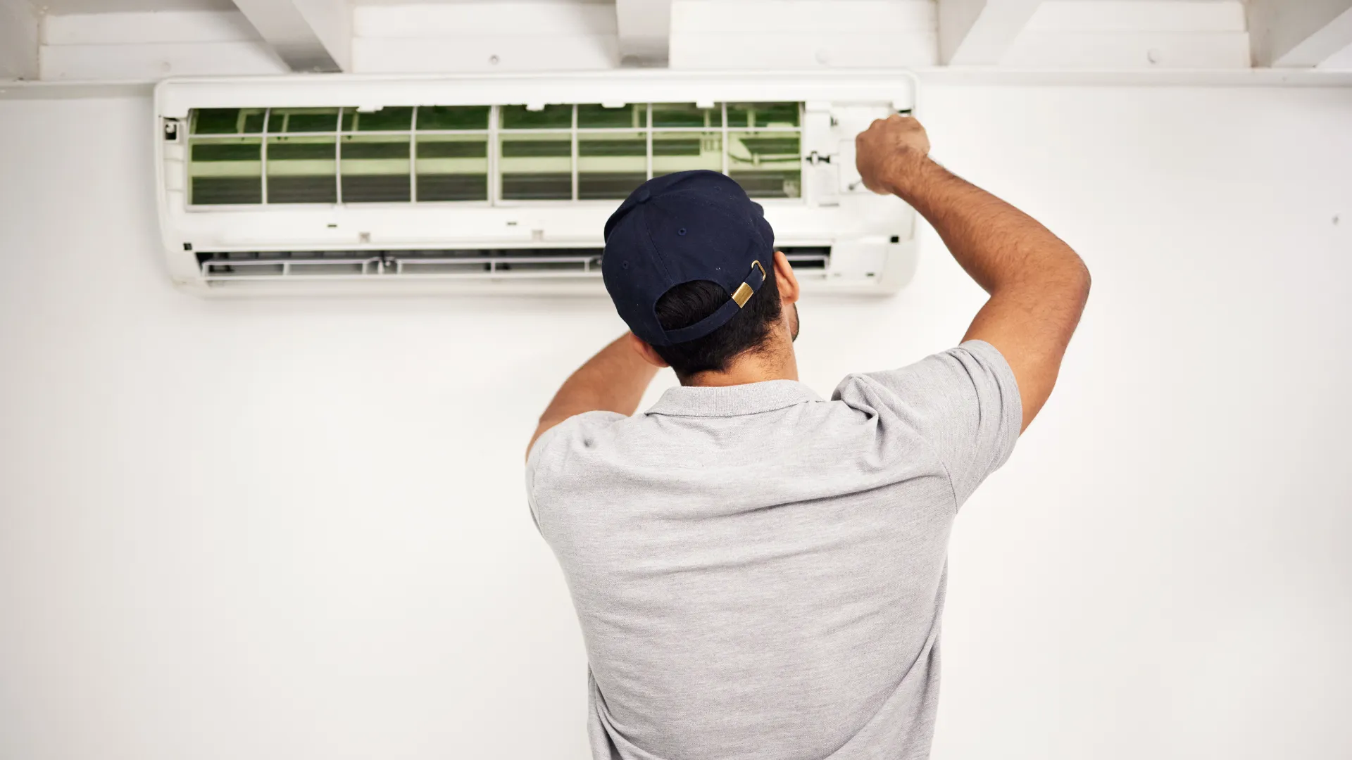 A person in a gray polo shirt and navy cap is servicing or repairing a wall-mounted air conditioner, with the cover open and tools in hand, facing the unit against a white wall.