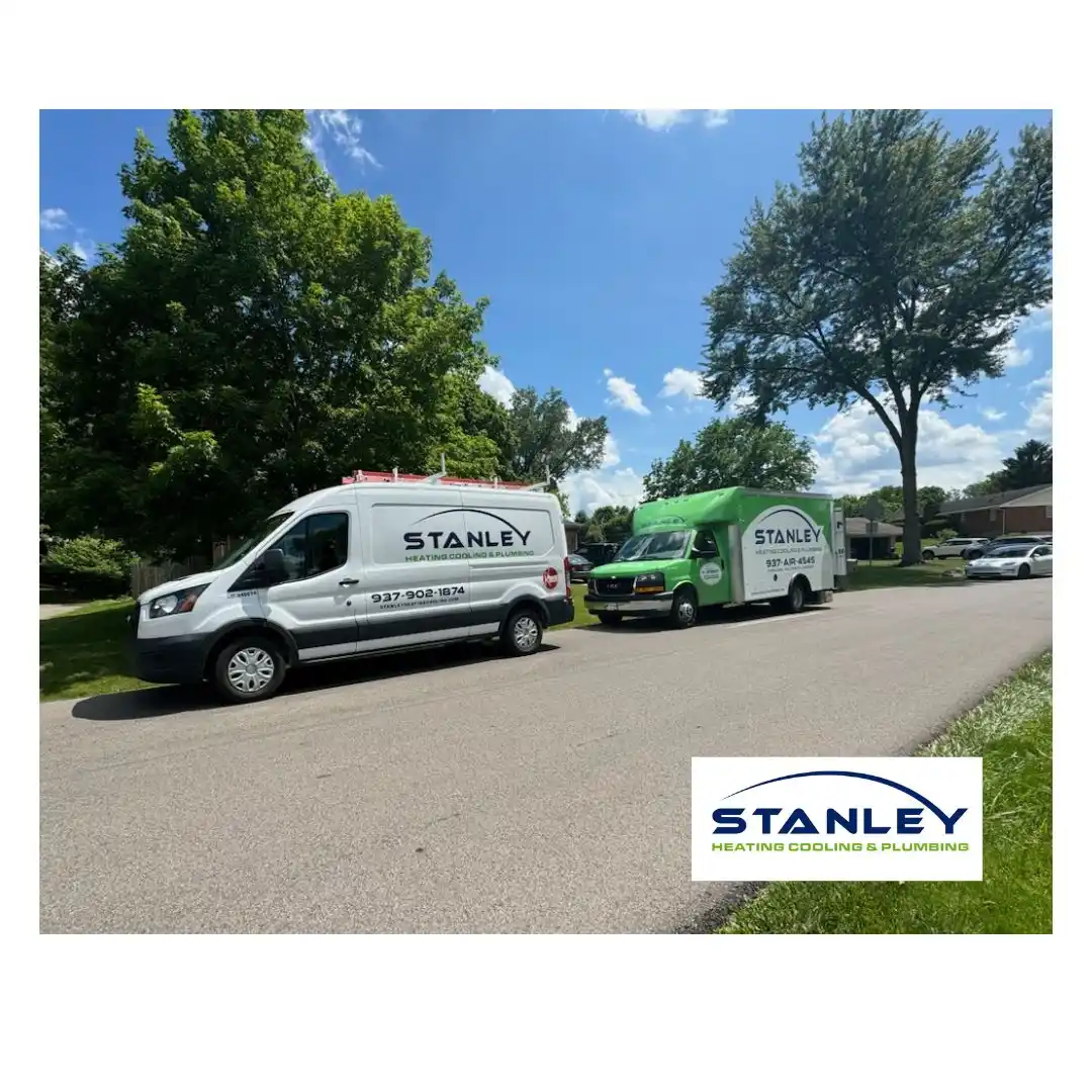 Two Stanley Heating, Cooling & Plumbing service vans, one white and one green, are parked on a suburban street with trees and houses in the background under a sunny sky—ready to help with any AC unit repair. The Stanley logo appears in the bottom right corner.