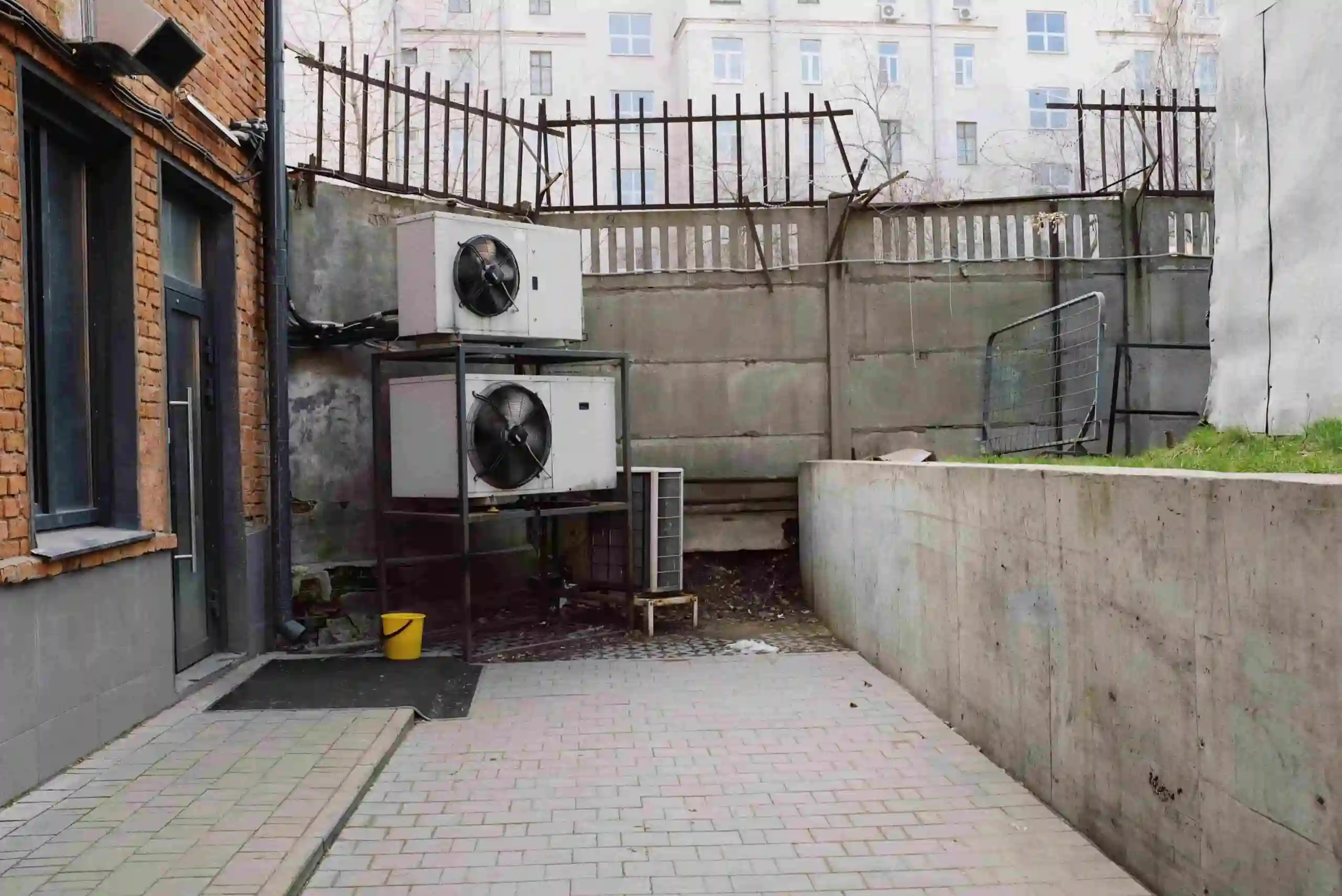 A small paved courtyard with two large air conditioning units, including an emergency HVAC system, mounted on a metal frame by a brick building, surrounded by high concrete walls and a metal fence on top. A yellow bucket sits near the wall.