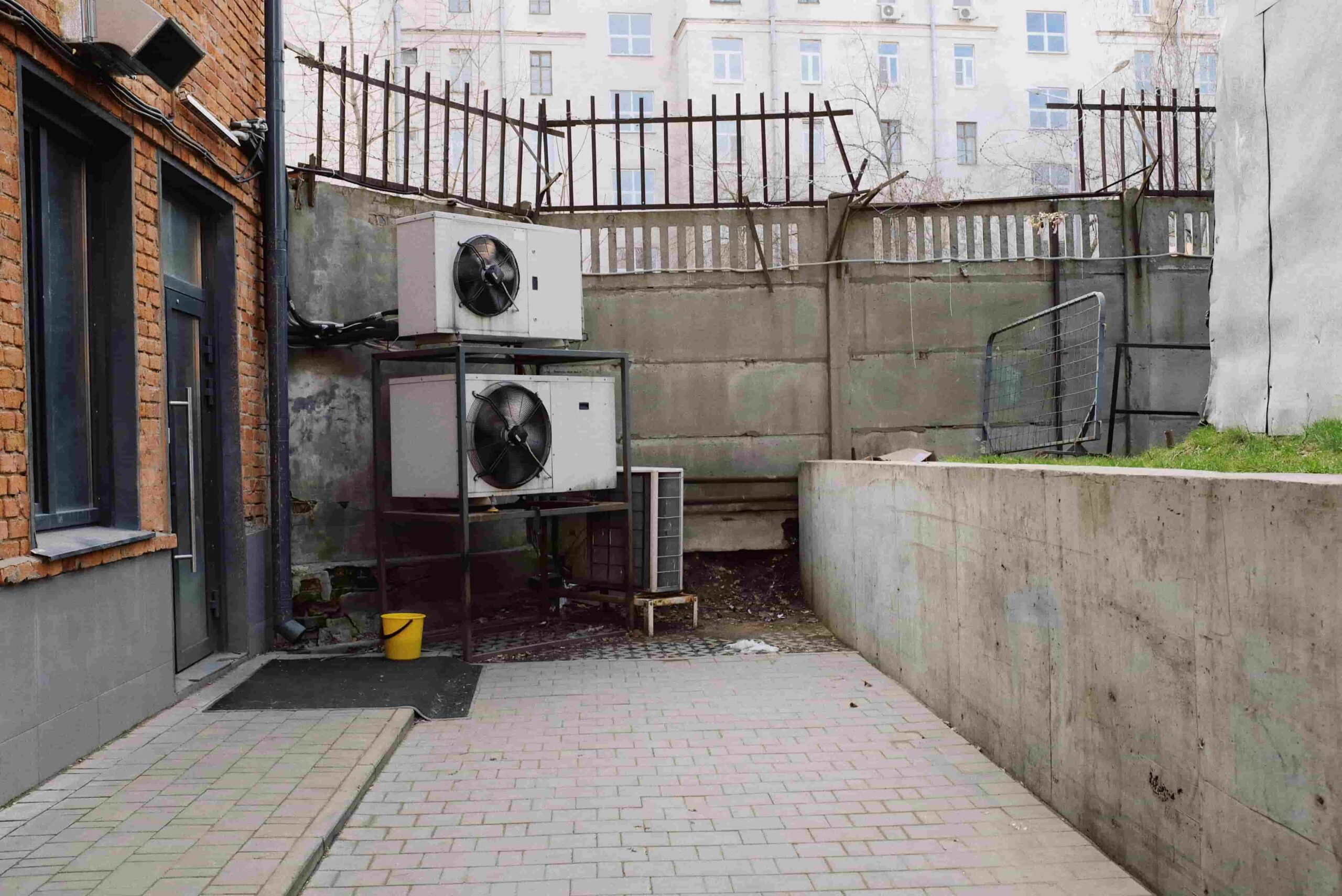 Two large air conditioning units are stacked in a small, walled alley with a tiled pathway and brick walls—ready for Emergency HVAC Dayton, OH: 24/7 Heating & Cooling Help. A yellow bucket sits nearby, with buildings visible in the background.