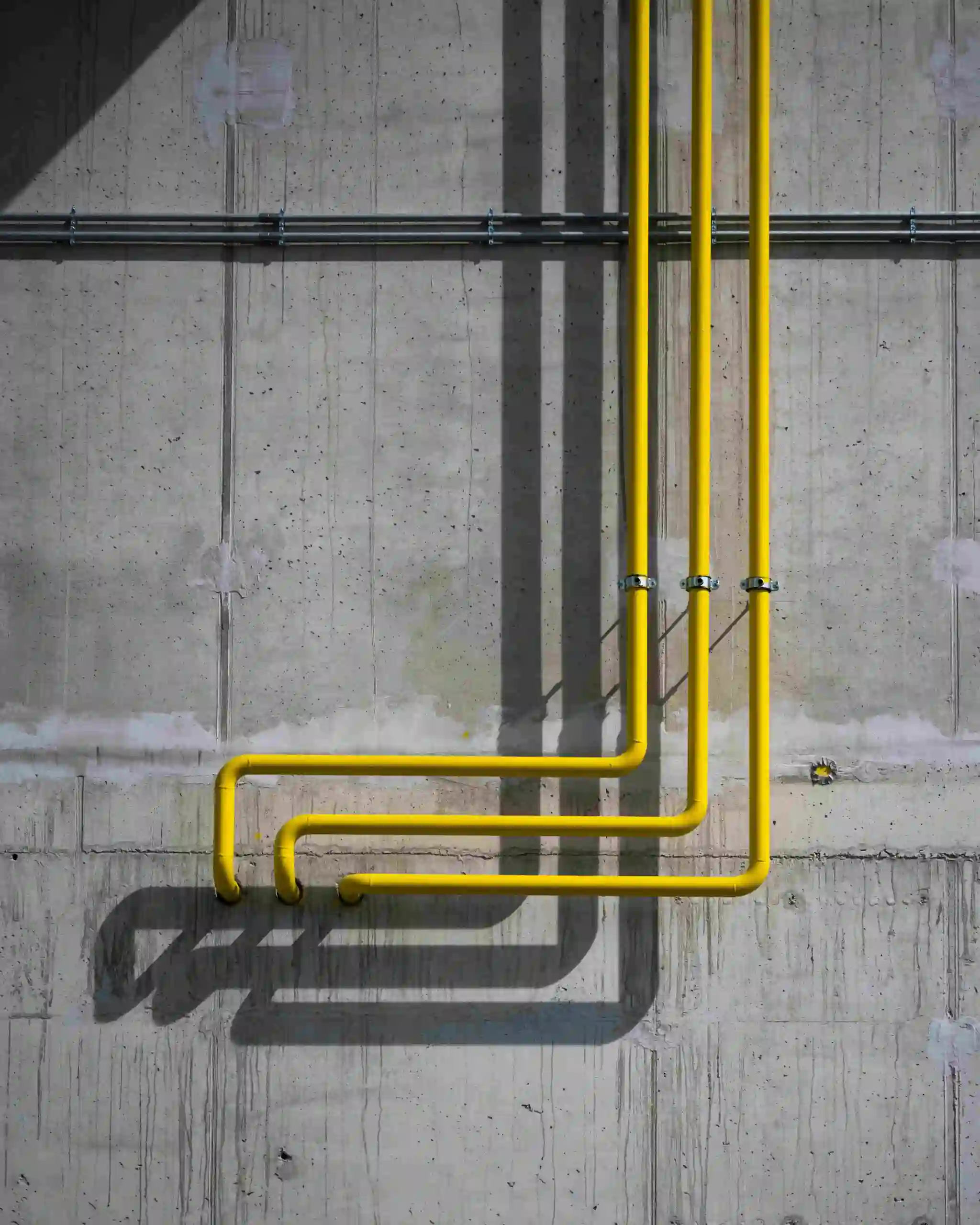 Three yellow pipes from a gas line installation run vertically down a gray concrete wall, then bend at right angles near the bottom, casting distinct shadows to the left. The wall shows some grime and texture.