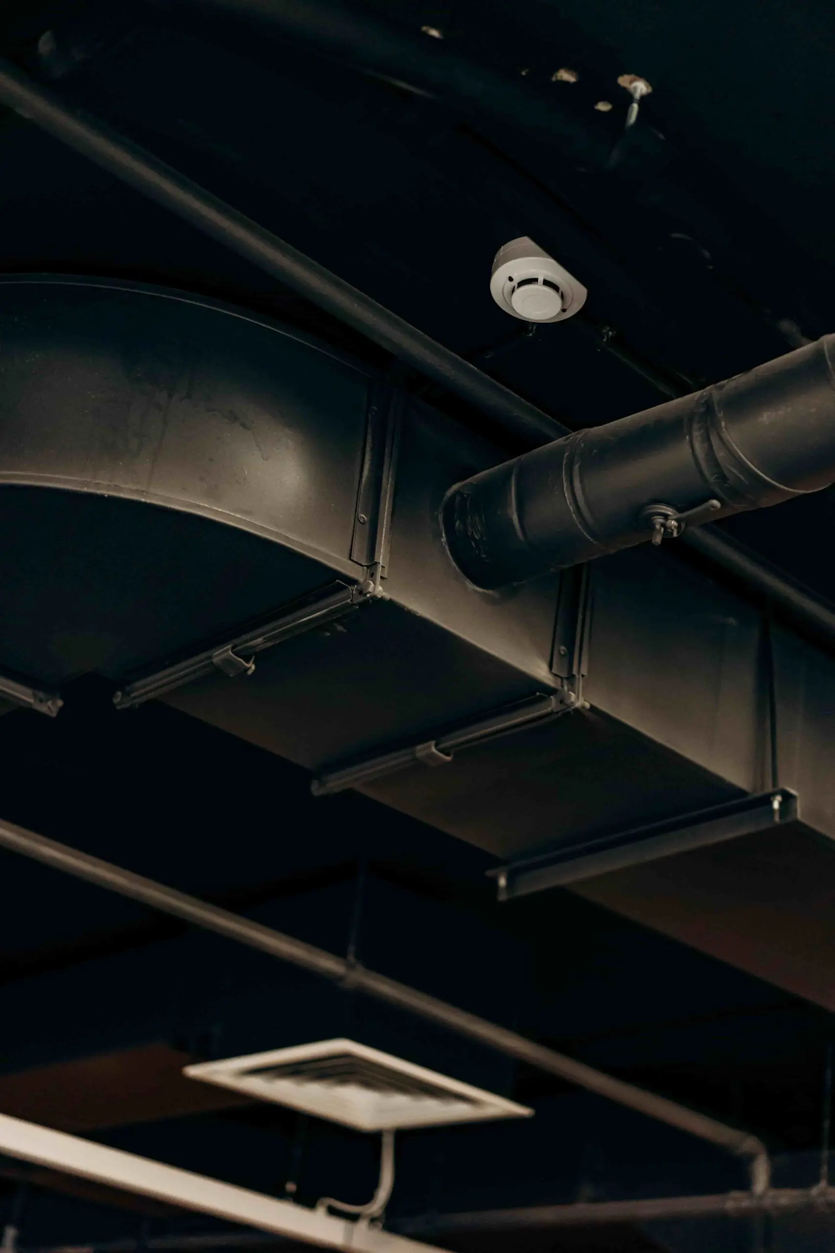 Exposed black industrial air ducts and pipes run along a dark ceiling, with a round smoke detector attached nearby—reflecting the quality installations seen with HVAC Service Dayton in commercial or industrial buildings.