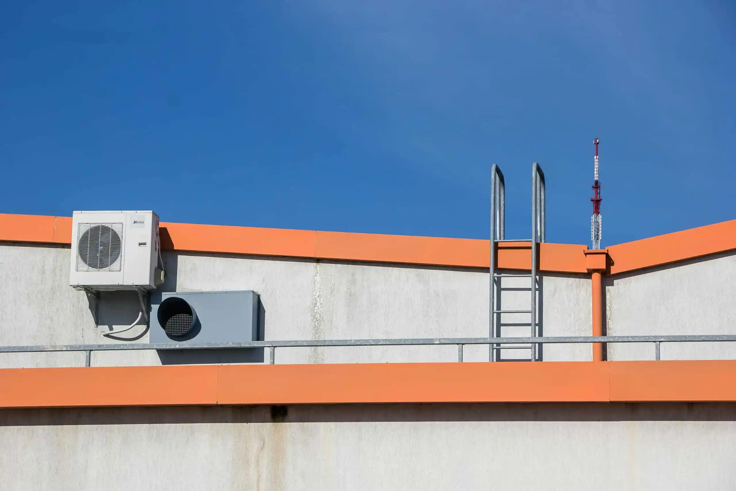 A metal ladder leads to the flat roof of a white building with orange trim, featuring Heating and Air Conditioning units and a vent against a clear blue sky.