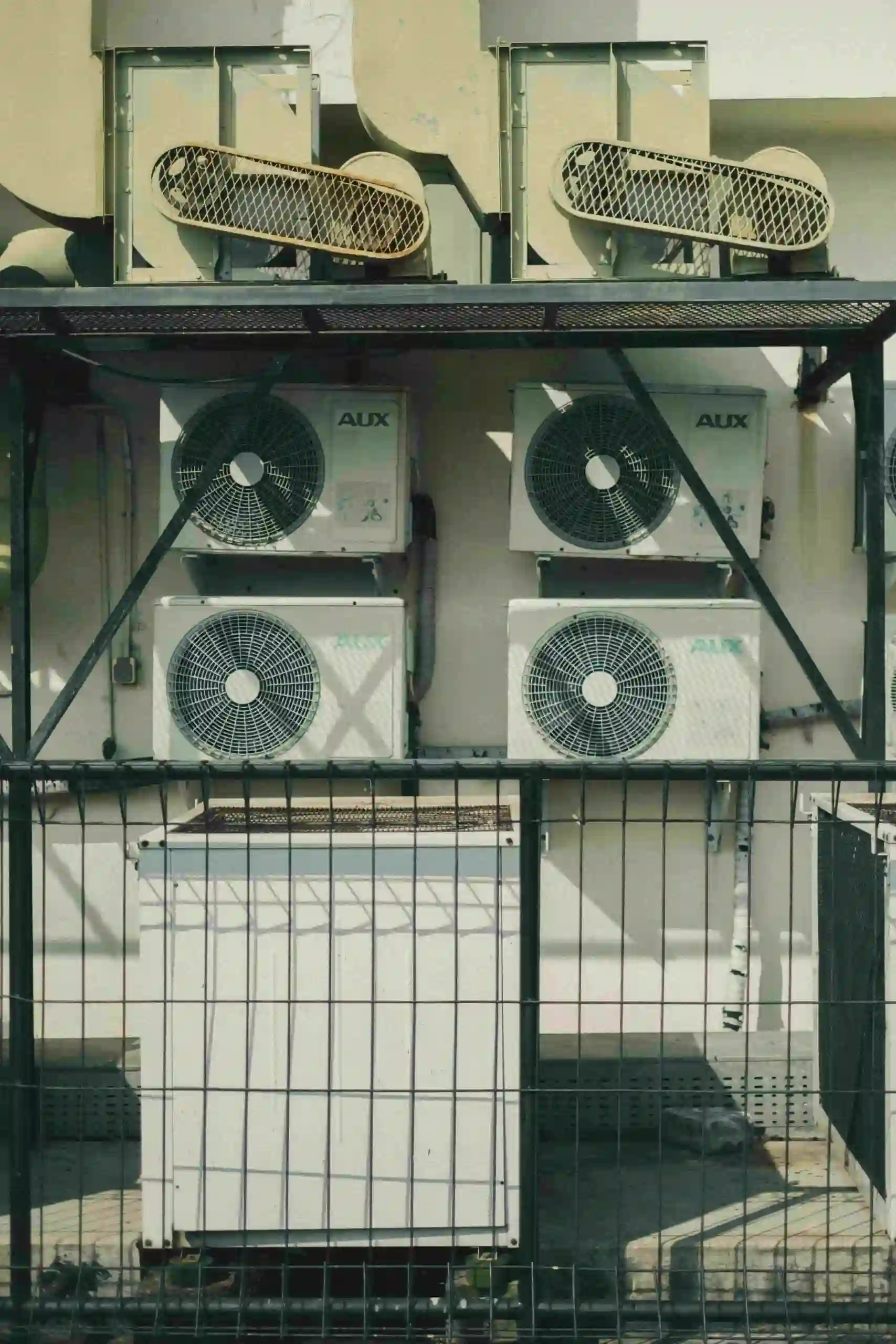 Four air conditioning units from Dayton Heating and Air are mounted on an exterior wall behind a black metal fence, with three additional yellowish units positioned on a metal platform above them. Sunlight casts shadows across the wall and units.