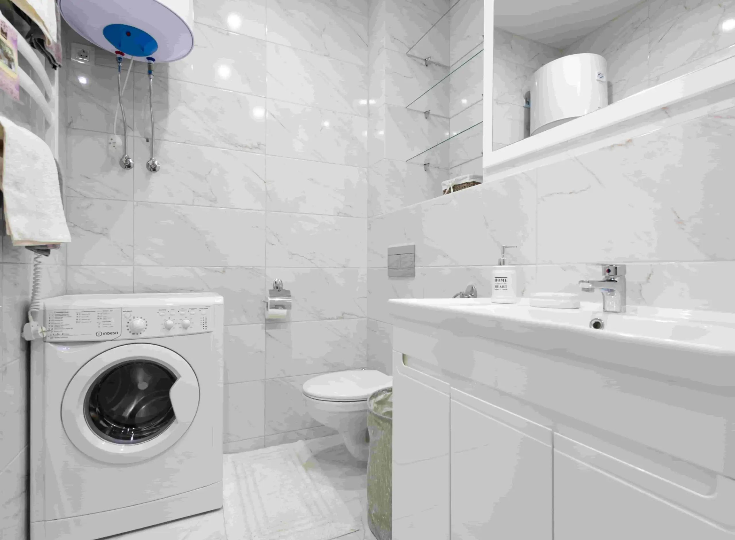 A modern, white-tiled bathroom features a washing machine, toilet, sink with cabinets, wall-mounted mirror, towel rack, and a sleek water heater. Shelves hold toiletries, maintaining a clean and organized atmosphere.