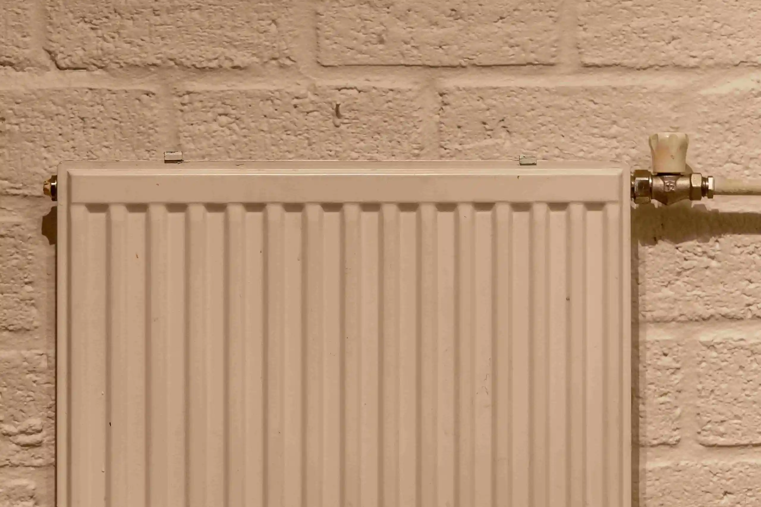 A close-up of a beige metal radiator, part of the heating and cooling system, mounted on a light-colored brick wall with a textured surface and a visible valve on the top right corner.