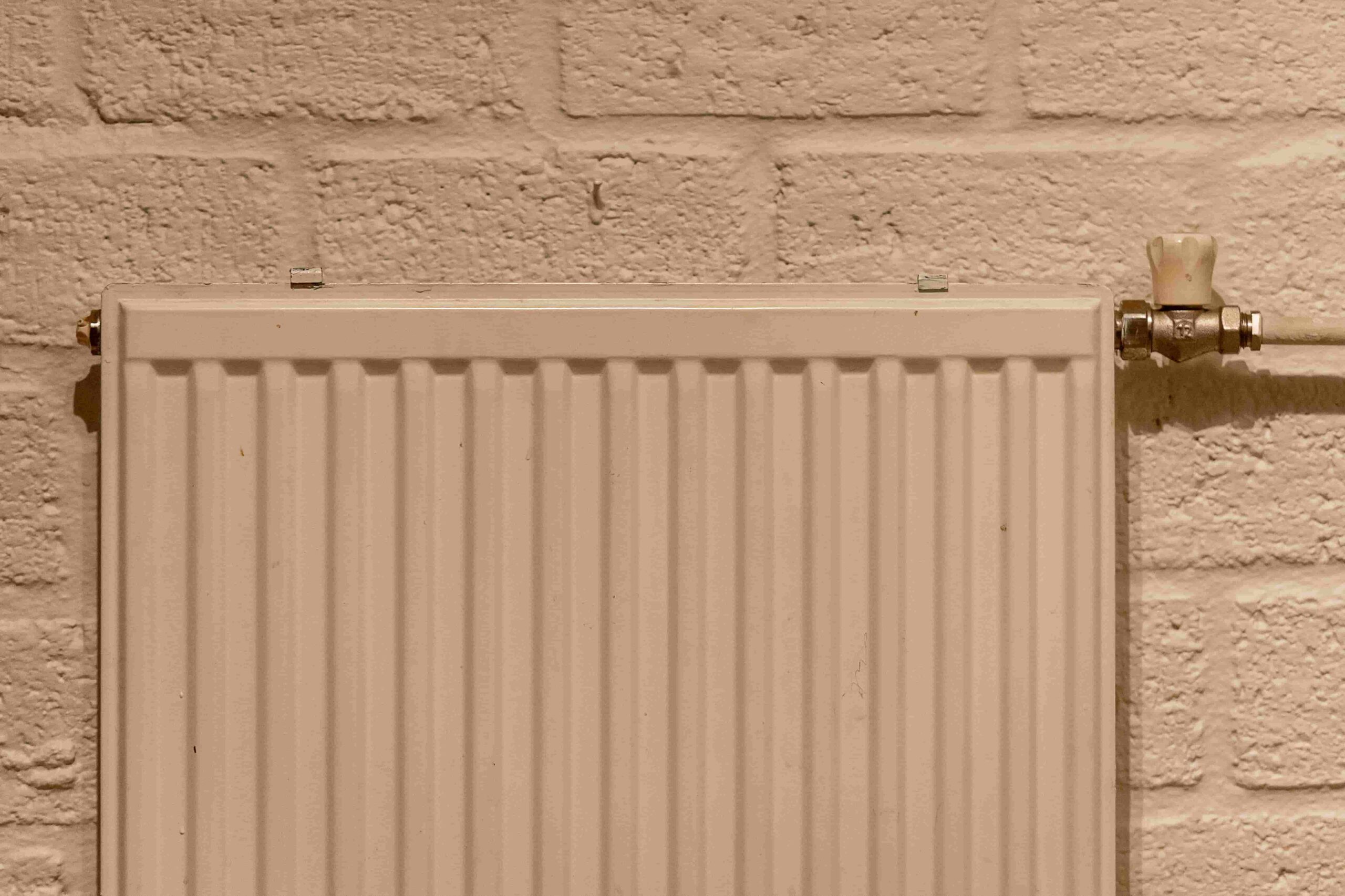 A white radiator with vertical ridges is mounted on a light-colored brick wall. The radiator's valve is visible on the upper right side, perfect for those seeking tips from The Ultimate Guide to Heating and Cooling in Dayton, OH.