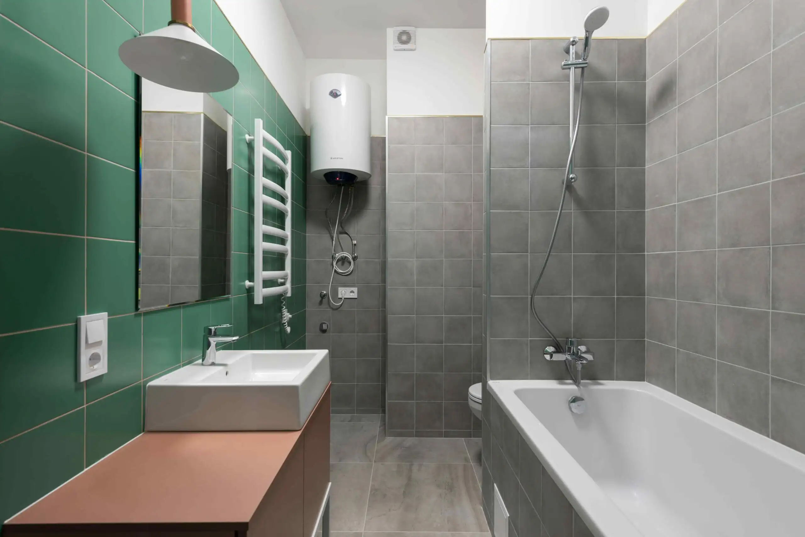 Modern bathroom with green and gray tiled walls, a white sink and cabinet, wall mirror, towel warmer, professional water heater installation, toilet, and a bathtub with a showerhead. The space is clean and minimalistic.