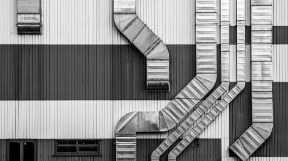 Black and white photo of a building’s exterior with large, angular metal ventilation ducts and pipes—similar to those installed by top HVAC companies—running vertically and diagonally across striped wall panels. Two small windows are visible at the bottom.