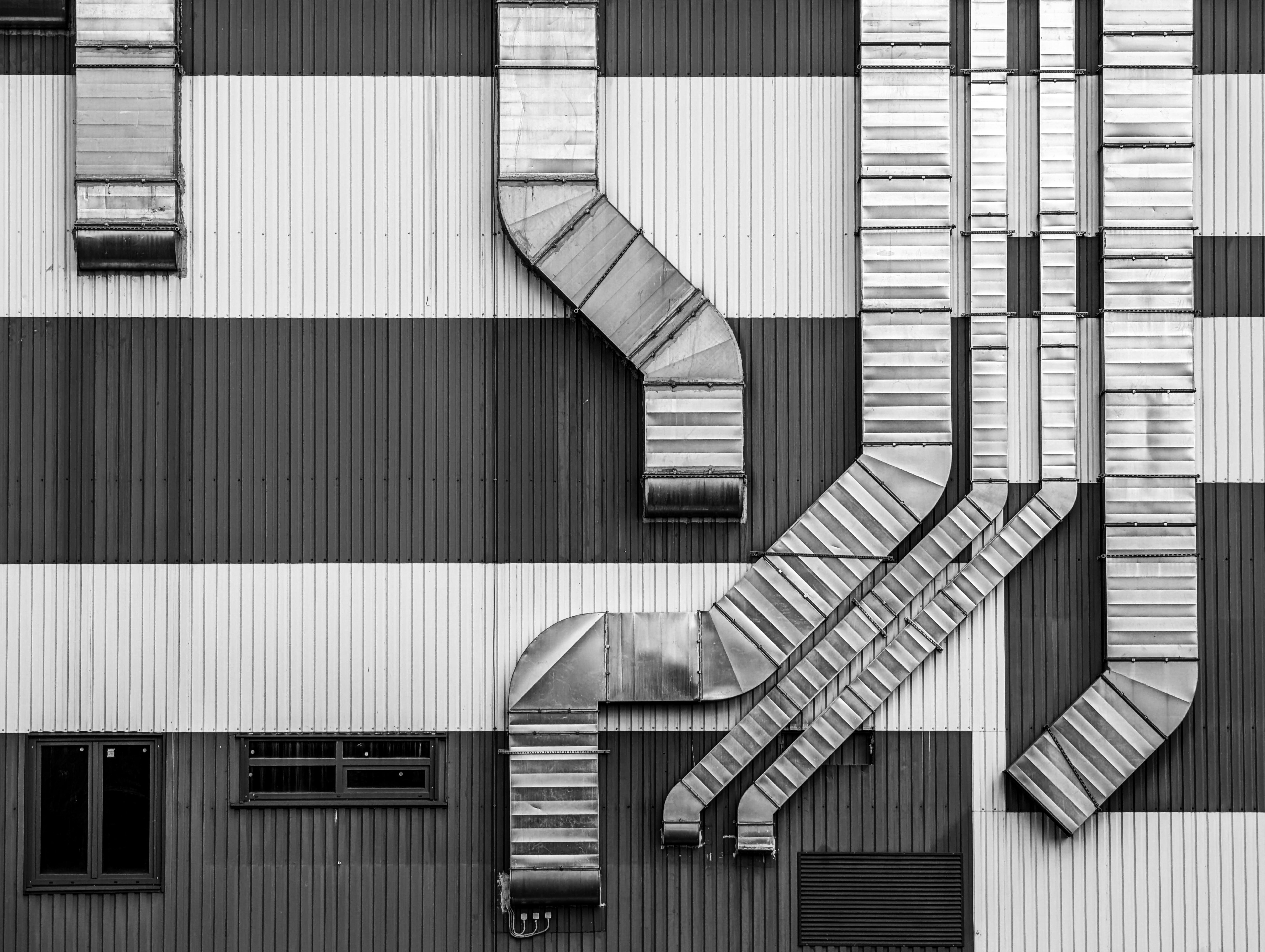 Black and white photo of a building’s exterior with bold horizontal stripes. Multiple metal ventilation ducts, hinting at Emergency HVAC Dayton services, run vertically and diagonally across the striped wall. A few small windows sit in the lower part.