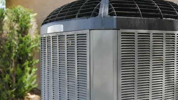 A close-up view of an outdoor air conditioning unit with metal vents and a protective grille on top, situated near a building and some greenery—ideal for illustrating Emergency HVAC Repair or HVAC Replacement Dayton services.