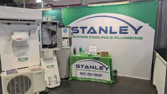 A trade show booth for Stanley Heating Cooling & Plumbing features a branded backdrop, HVAC equipment display, informational brochures, and a green table with company signage, contact information, and details about Free Estimates—one of the leading HVAC companies Dayton.