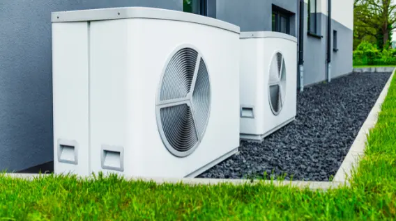 Two modern outdoor heat pump units are installed next to a building on dark gravel, with green grass in the foreground. For professional heat pump installation or quotes in Dayton OH, contact us today.