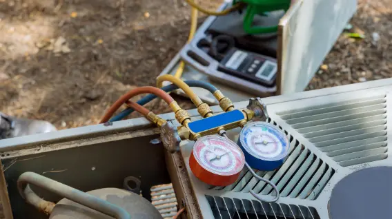Close-up of HVAC manifold gauges attached to an outdoor air conditioning unit in Dayton, showcasing Affordable HVAC Repair tools and equipment on a dirt surface in the background.