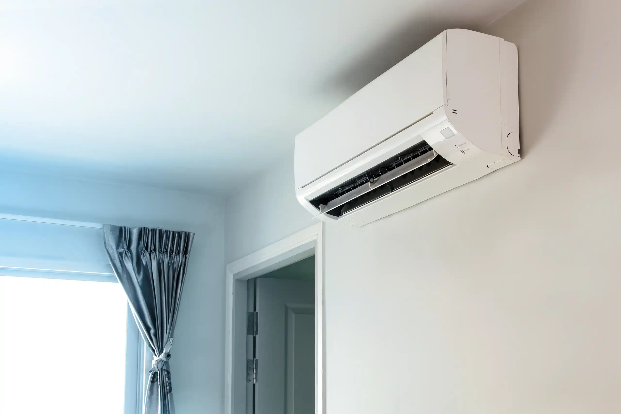 A white wall-mounted air conditioner, reminiscent of the efficient Dayton air conditioning units, is installed above a doorway in a bright room with light walls, a door, and a window covered by a gray curtain tied to the side.