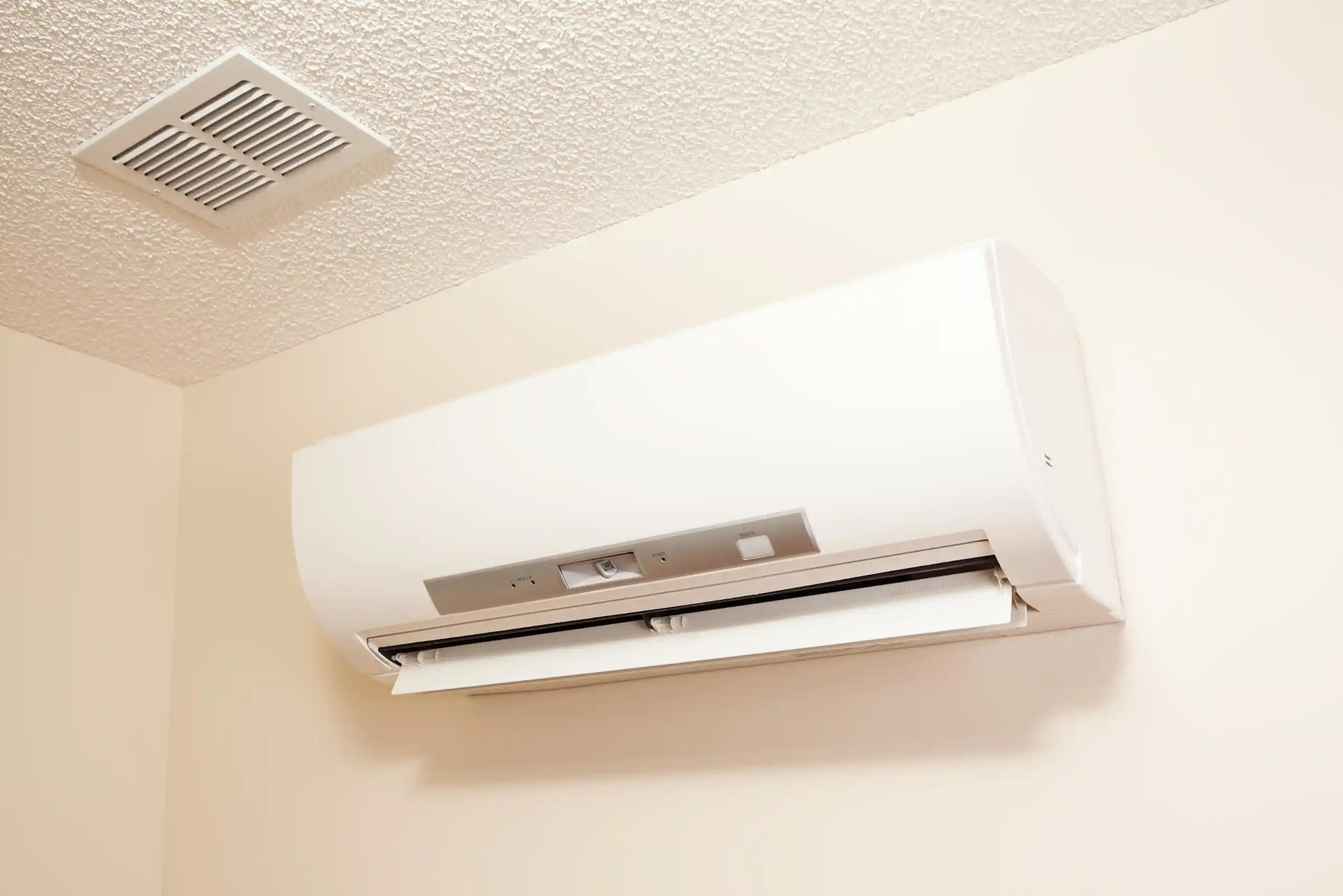 A white wall-mounted ductless mini-split air conditioning unit is installed near the ceiling of a beige room in Ohio, with a vent visible nearby on the textured ceiling—showcasing why choosing ductless mini-splits truly is worth it for efficient climate control.