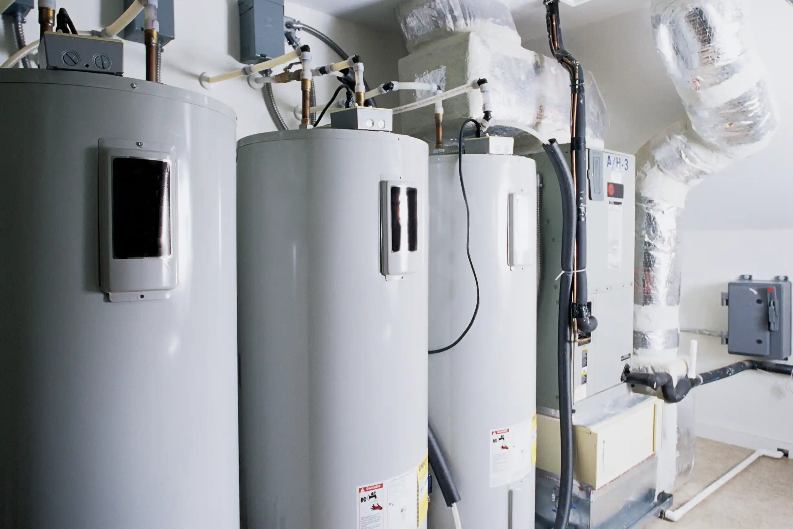 Three large water heaters and an HVAC unit are installed in a utility room, with various pipes, ducts, and electrical connections visible. The room has bright lighting and white walls. When considering how long water heaters last, it’s important to note that the water heater life expectancy can vary depending on usage and maintenance.