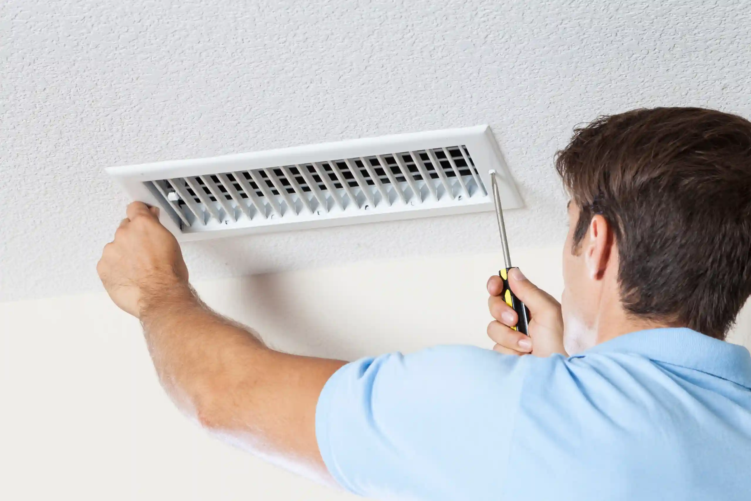 A man in a light blue shirt uses a screwdriver to remove or install a vent cover on a white ceiling, perhaps as part of routine home maintenance similar to checking on your water heater lifespan and ensuring water heater durability.