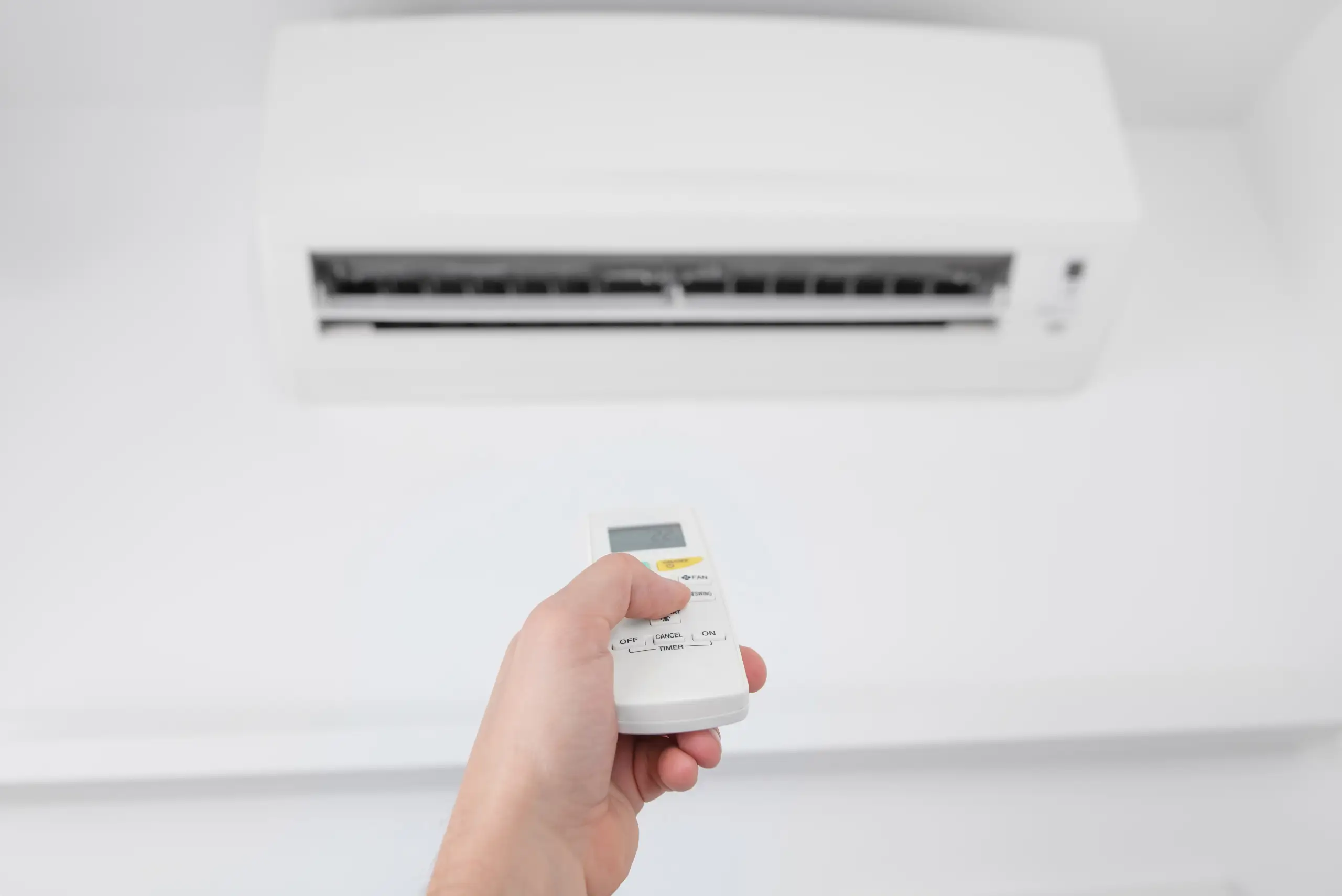 A person’s hand holding a remote control and pointing it at a white wall-mounted air conditioner unit, possibly checking for air conditioner problems or performing some AC troubleshooting due to the AC not cooling properly.